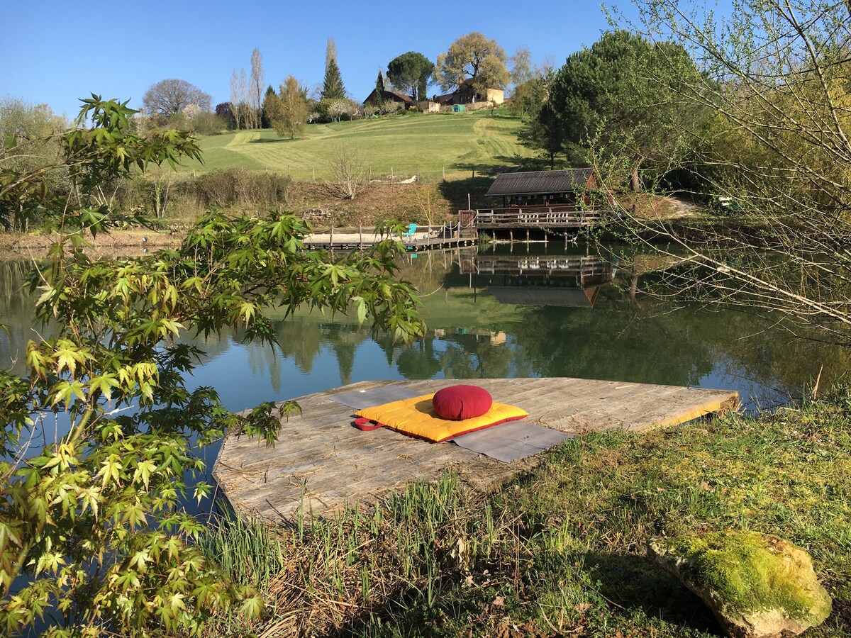 A serene wooden platform extends over calm waters, featuring a meditation mat and cushions. Lush greenery frames the scene, with reflections of trees and structures visible in the water. In the background, a rustic cabin and additional decking are nestled among the trees.