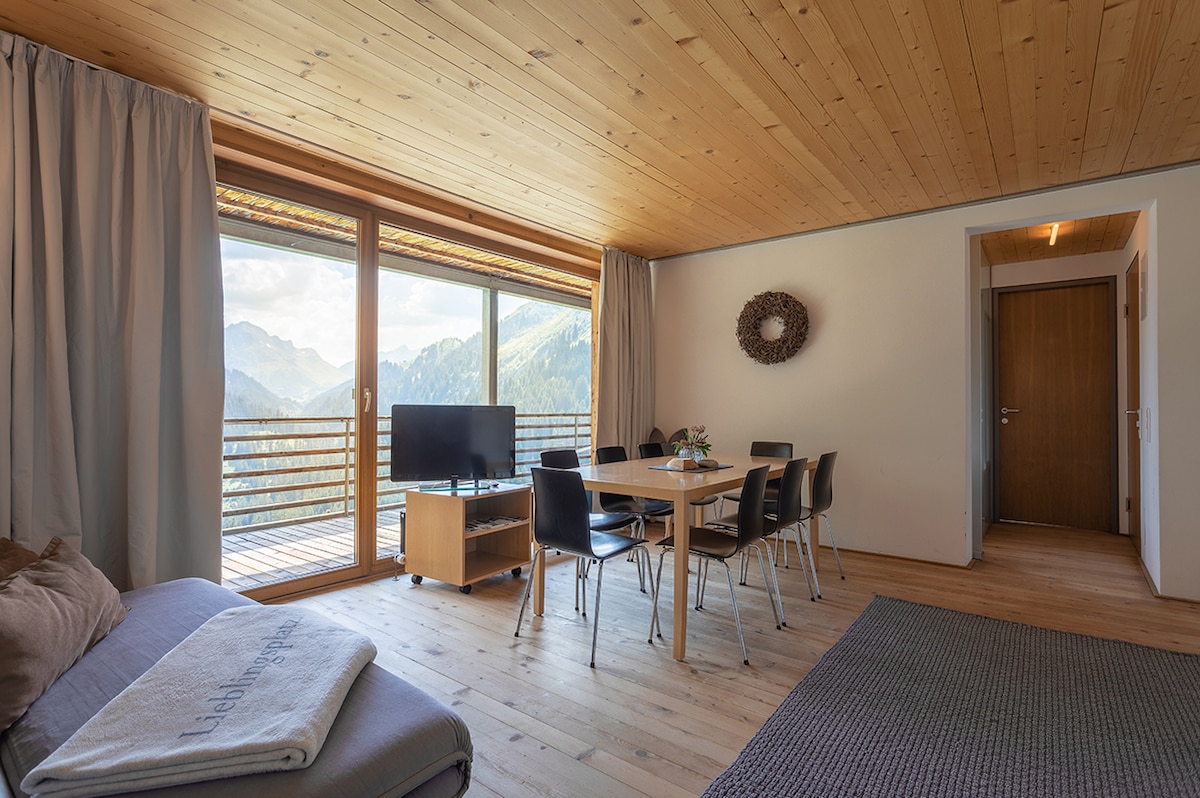 The spacious living area features a large table surrounded by black chairs, with a television positioned near the large windows. Natural light floods the room, highlighting the wooden ceiling and floor. A balcony provides stunning mountain views, enhancing the overall atmosphere.