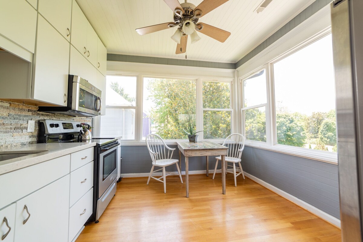 The bright kitchen features large windows on three walls, providing ample natural light and views of greenery. A small dining table with two white chairs is situated near the windows, accompanied by stainless steel appliances and light-colored cabinetry.