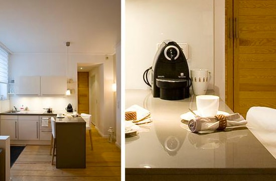 A modern kitchen features light cabinetry and a spacious island with seating. Stainless steel appliances and a sleek countertop complement the inviting atmosphere. A coffee machine is positioned next to dishware, providing a functional and stylish workspace adorned with neatly arranged linens.