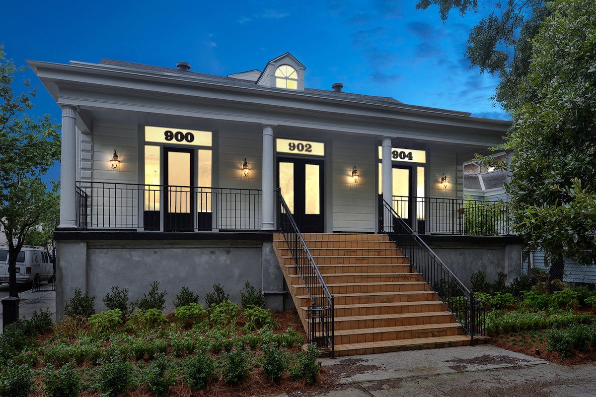 A charming exterior view of a multi-unit building is presented, featuring three distinctive entrances numbered 900, 902, and 904. Steps lead up to the front doors, framed by large windows on either side, illuminating the welcoming atmosphere. Lush greenery surrounds the base of the structure.