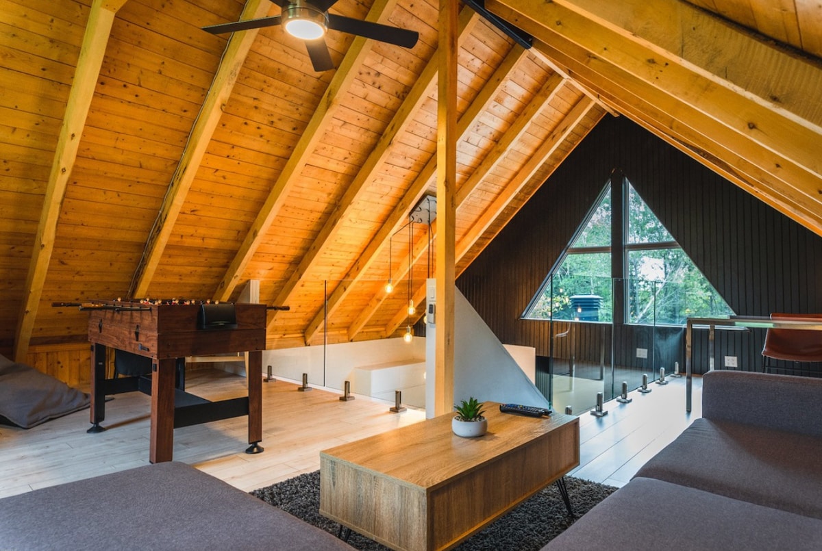 An open mezzanine lounge area features a sectional sofa and a wooden coffee table. A foosball table occupies one corner, while windows offer views of the surrounding nature. High wooden ceilings and exposed beams accentuate the space.