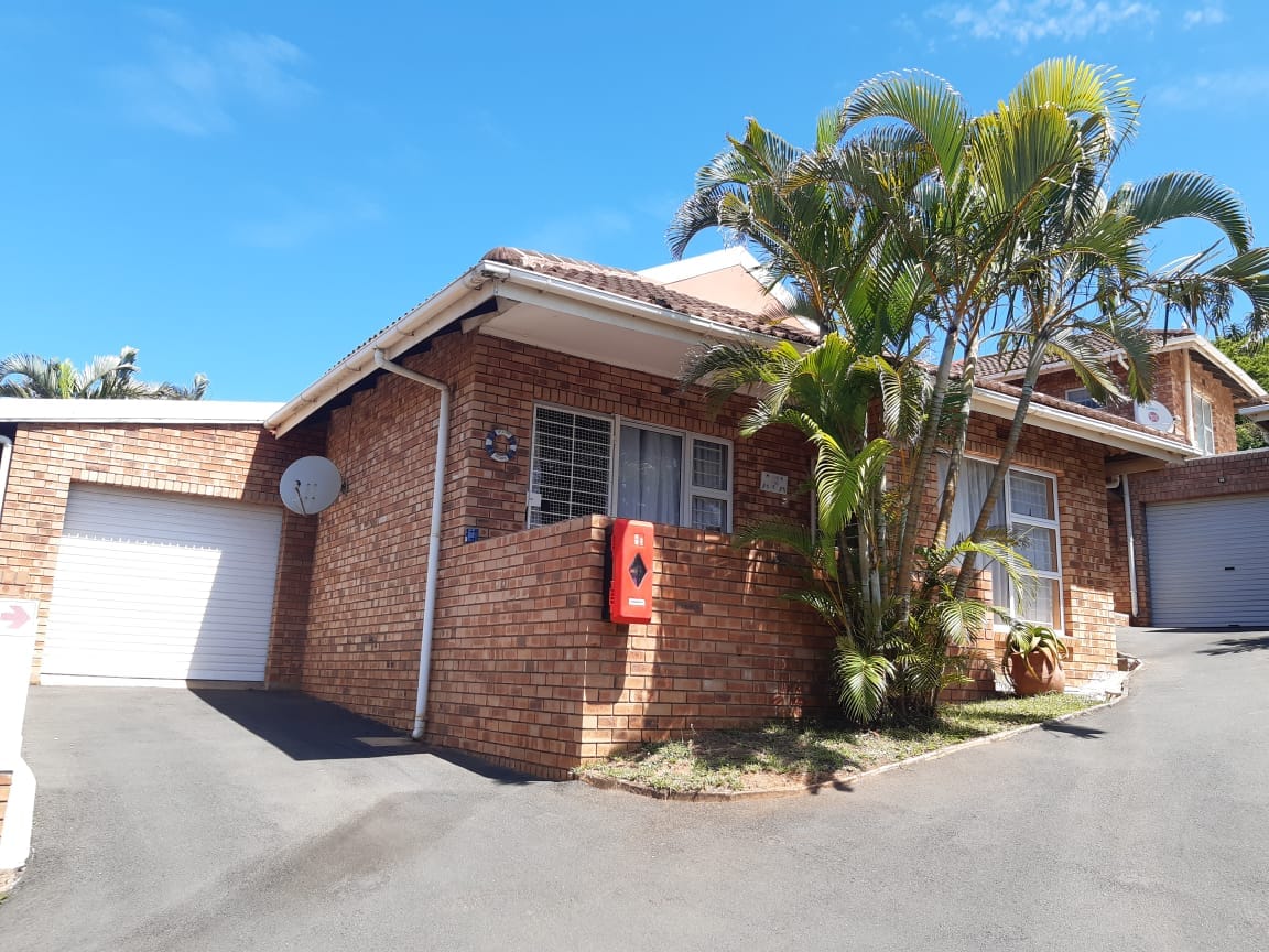 A brick exterior of a standalone unit is set against a clear blue sky. Lush green palm trees are positioned near the entrance, while the driveway leads to a garage. Windows are framed by light curtains, enhancing the home's welcoming appearance.