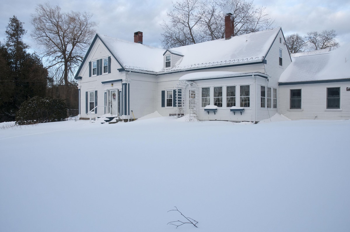 A historic 1800's shipbuilder's home is set against a snowy landscape, showcasing a white exterior with blue accents. Large windows are visible, allowing natural light to brighten the interior. Snow blankets the ground, creating a serene and quiet atmosphere around the residence.