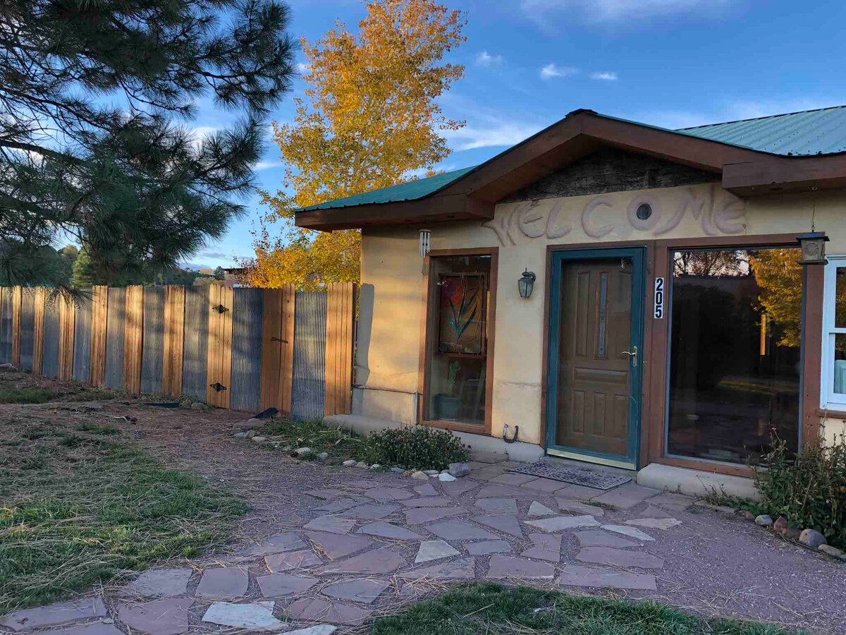 The exterior of the Strawbale home features a welcoming entrance adorned with the word 'WELCOME' carved above the door. A stone pathway leads to the door, bordered by landscaped areas. The property is enclosed by a wooden fence, with autumn foliage visible in the background.
