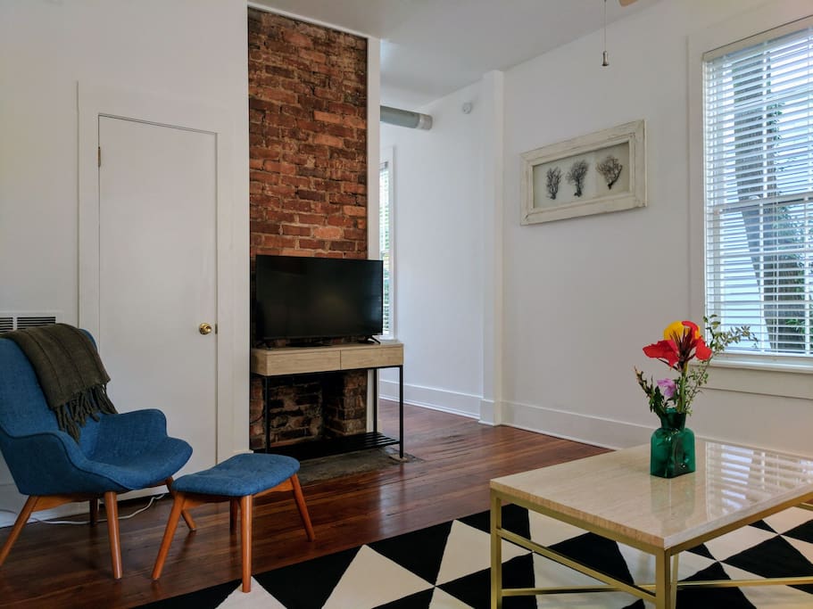 A view of the living room looking in from the front door entrance. The original, decorative brick fireplace has been restored. Two large wooden windows let tons of sunshine into the space.