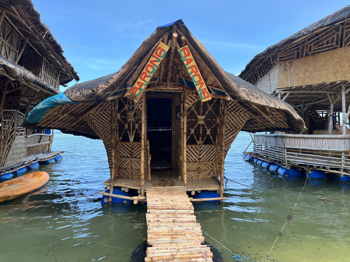 A floating cabin features a distinctive roof design, made of bamboo and thatch. A bamboo walkway connects the cabin to the surrounding water, with shades of blue reflecting off the surface. The entrance is marked with a sign that reads 'BARONG.'
