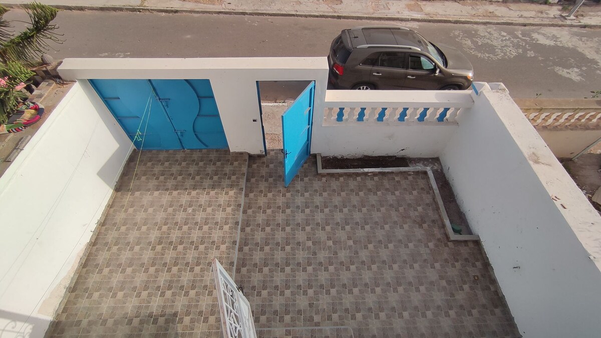 An outdoor area is visible, featuring a paved floor with a geometric pattern. A bright blue gate is shown partially open, leading to a clear path. A parked vehicle is seen outside the premises, indicating accessibility to the street.