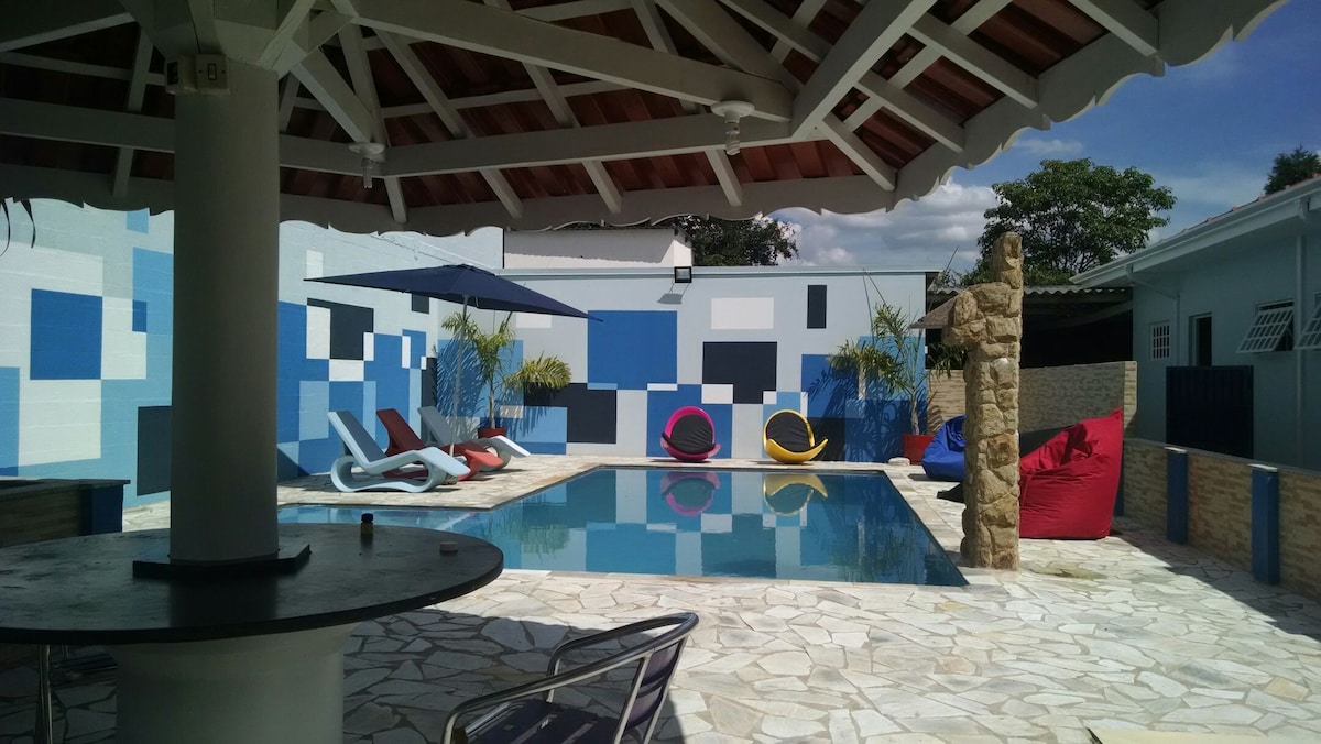 A vibrant outdoor area includes a swimming pool surrounded by colorful seating options. Poolside, a shaded area with a ceiling fan provides comfort. Surrounding walls feature abstract geometric patterns, and lush plants offer a natural touch to the space.