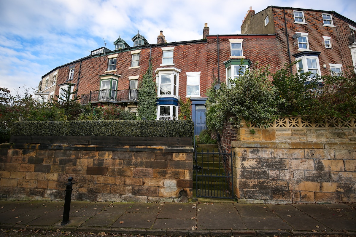 The Victorian town house features red brick and stonework, showcasing multiple levels with balconies. Lush greenery frames the entrance, which is accessed by a set of steps leading through an iron gate. Windows with decorative trim provide character to the facade.