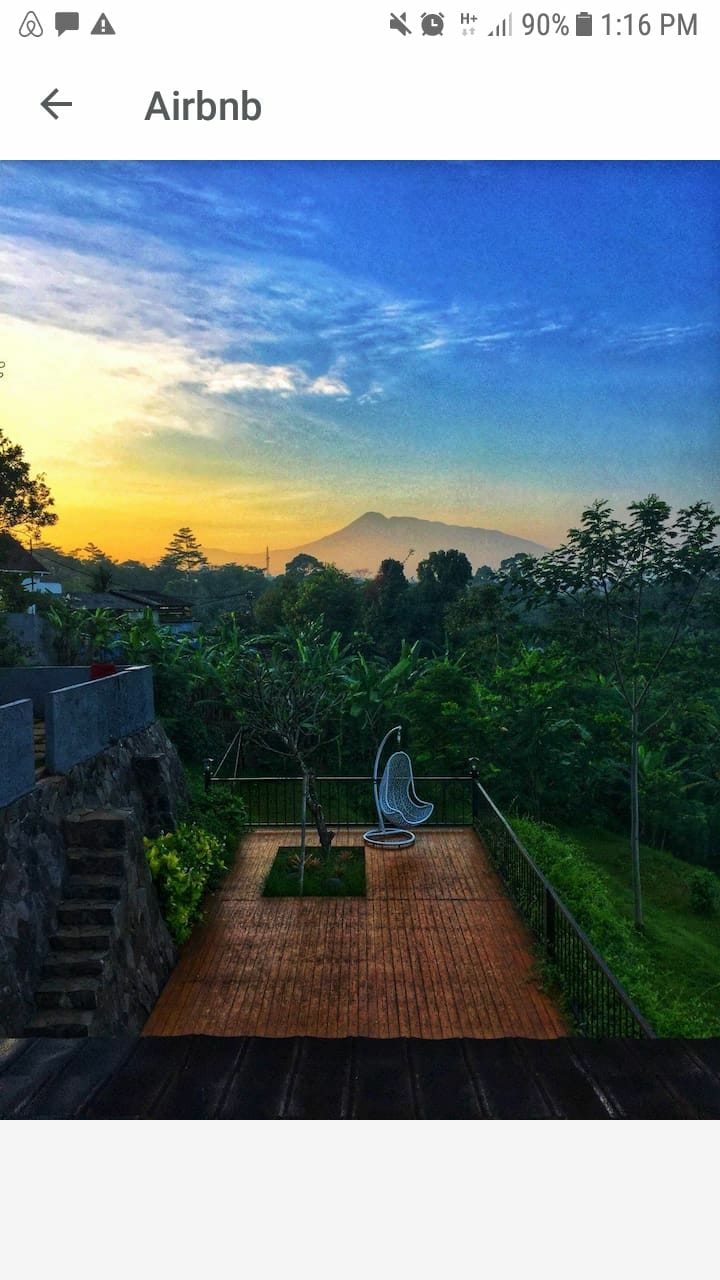 A wooden deck is situated in a lush green setting, featuring a white hanging chair. The backdrop showcases distant mountains during sunset, with soft colors in the sky. Surrounding greenery adds to the serene atmosphere.