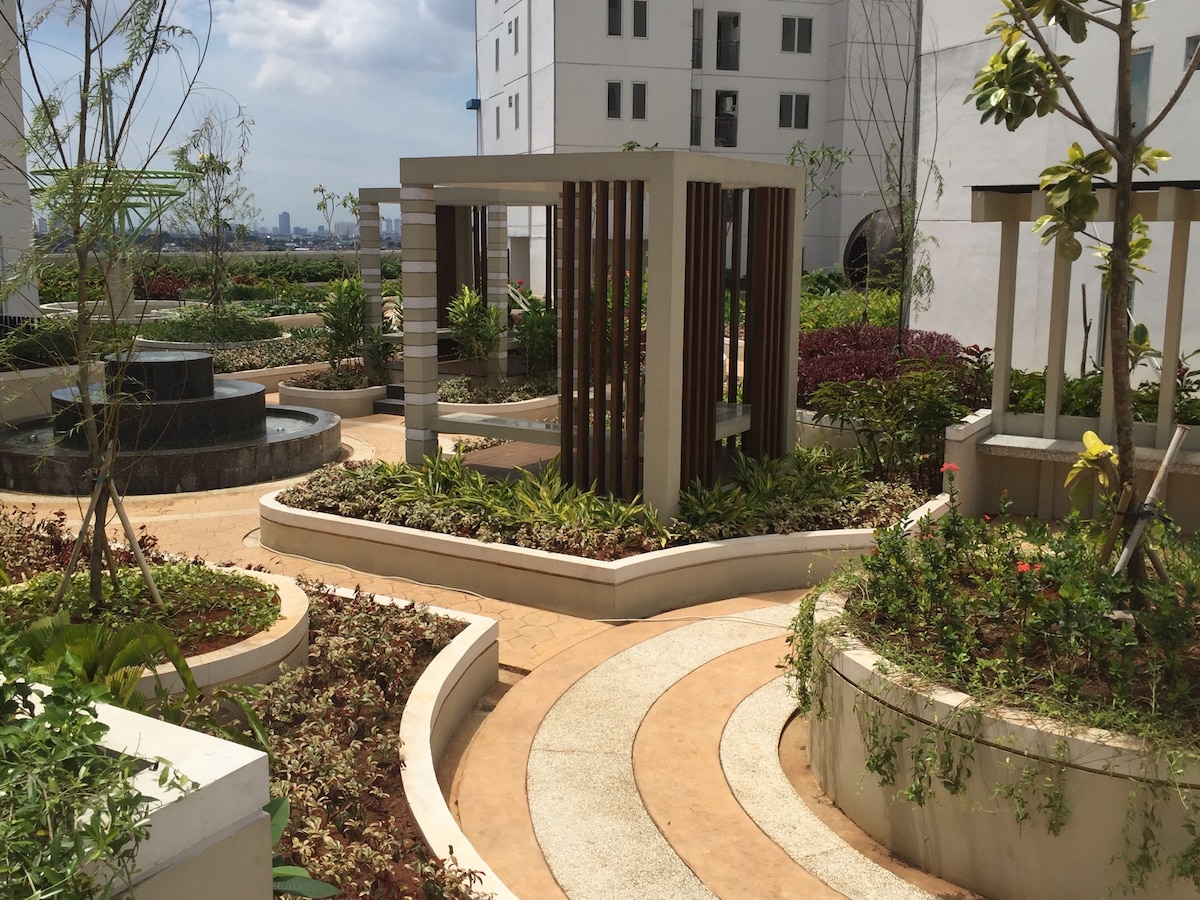 A landscaped garden area is showcased, featuring winding pathways surrounded by colorful plants and shrubs. Several pavilions with wooden accents are situated throughout the space, alongside a small fountain. The surroundings offer a serene atmosphere for relaxation amidst greenery.