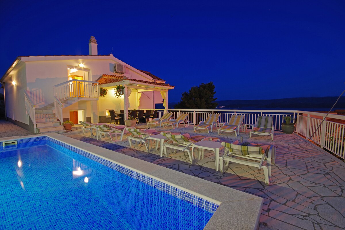 A spacious villa exterior is depicted at dusk, featuring a terrace with multiple lounge chairs arranged around a swimming pool. Soft lighting highlights the architecture and creates a serene ambiance, with a clear sky and distant views enhancing the tranquil setting.