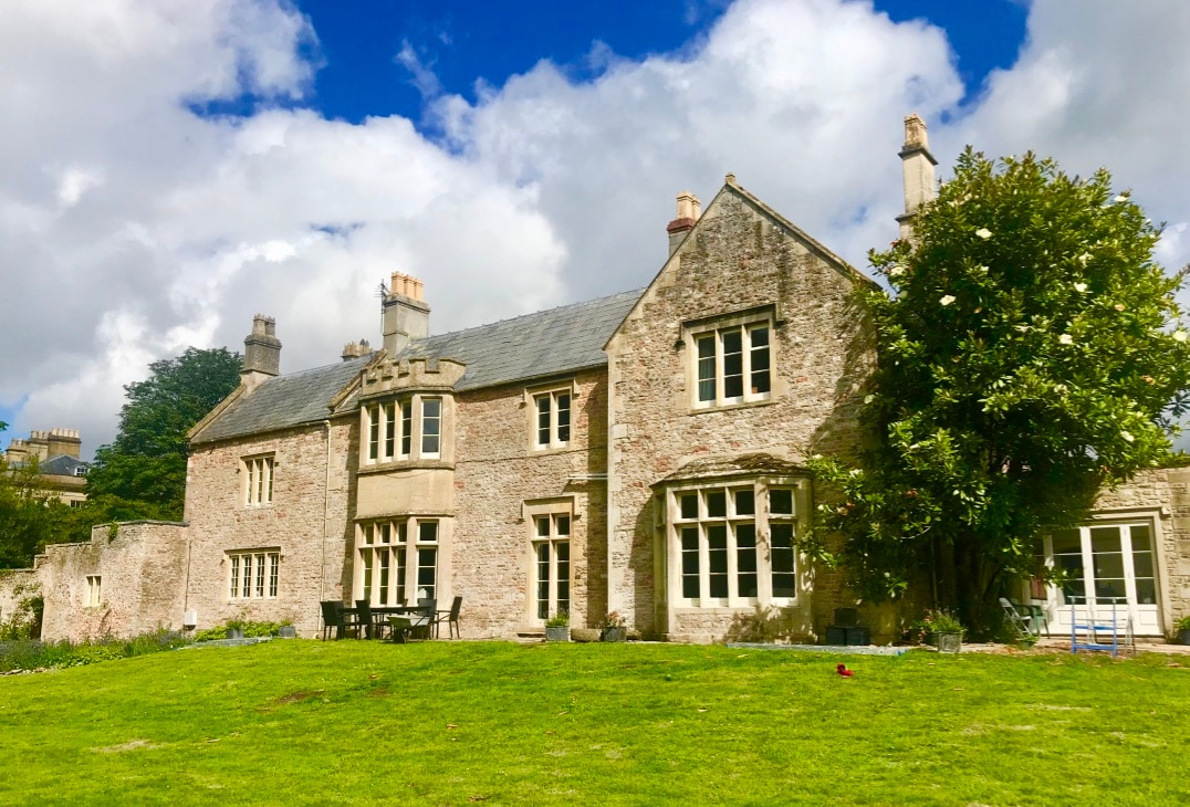 The West House is a stone structure featuring large windows and a mix of architectural styles. A private walled garden surrounds the property, showcasing a lawn area planted with various greenery. The sky is partly cloudy, enhancing the serene countryside setting.