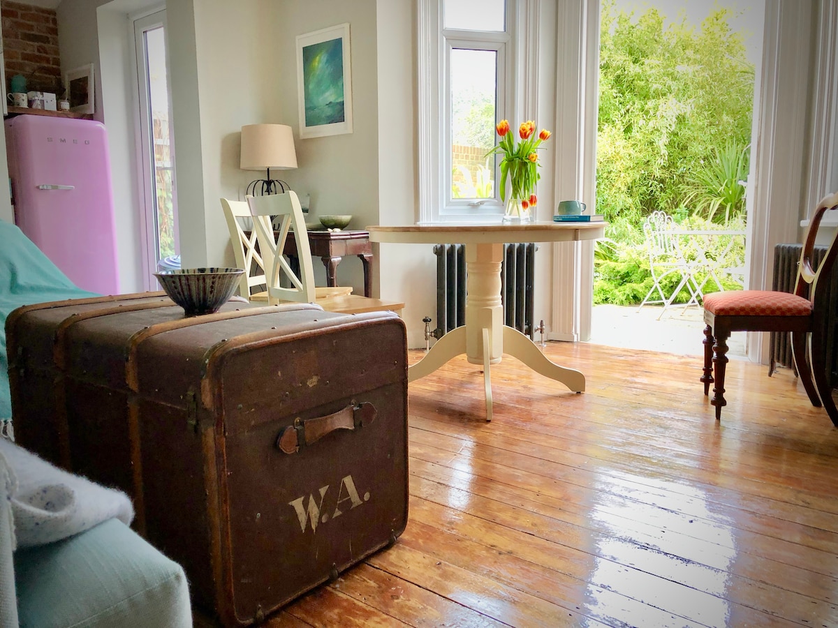 A light-filled dining area features a round table surrounded by simple chairs. A vintage trunk is positioned nearby, and a vase with tulips adds a pop of color. Large windows provide views of a garden with greenery, enhancing the inviting ambiance.