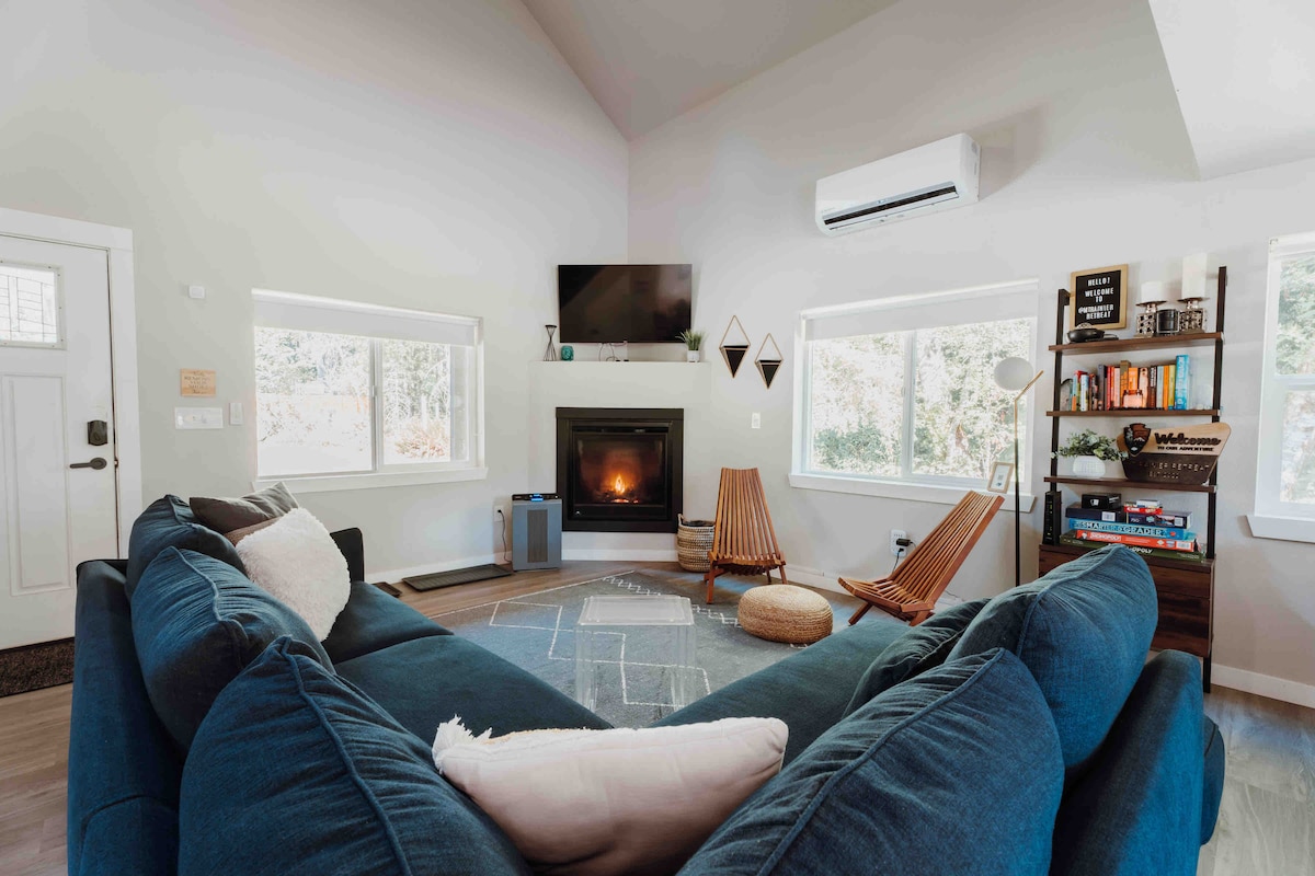 A spacious living area features a large blue sectional sofa, complemented by two wooden chairs and a round ottoman. Natural light filters through window panels on either side of a modern fireplace, while a mounted television offers entertainment options.