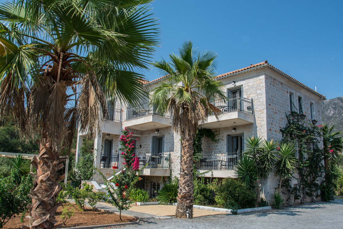 A three-story stone building is surrounded by tropical palm trees and lush greenery. Colorful flowers add vibrancy to the landscape, while balconies off the upper levels overlook the garden. The clear blue sky complements the serene outdoor environment.