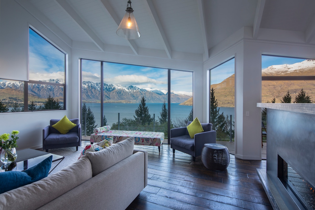 A contemporary living area features floor-to-ceiling glass windows on three sides, providing panoramic views of Lake Wakatipu and surrounding mountains. A plush sofa sits adjacent to two armchairs. A coffee table with decorative accents occupies the center, while a fireplace adds warmth and character.