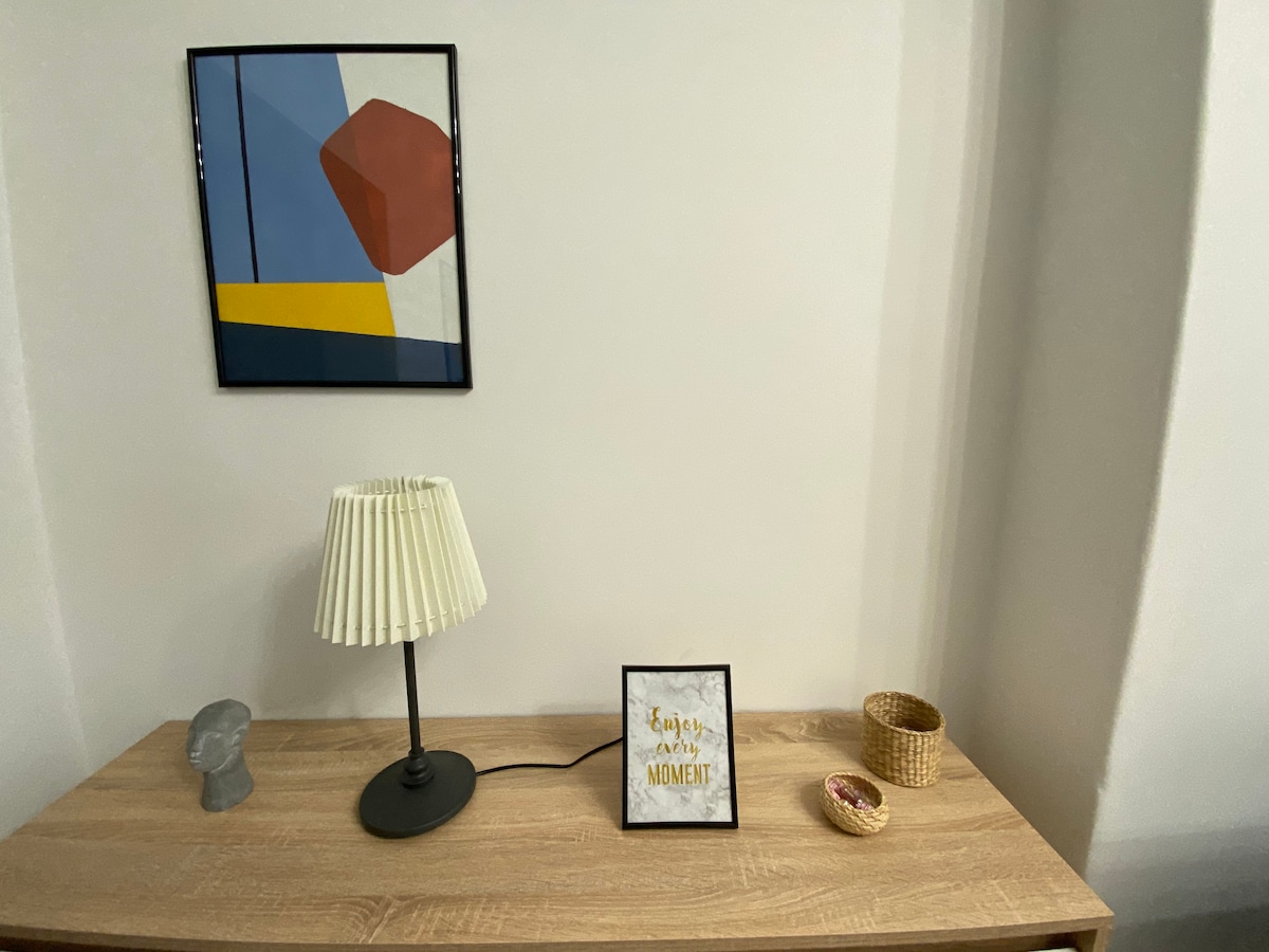 A wooden surface displays a variety of decorative items, including a small sculpture, a lamp with a pleated shade, and a framed quote. A woven basket is positioned to the right, with a colorful trinket resting inside. A geometric artwork adorns the wall above.