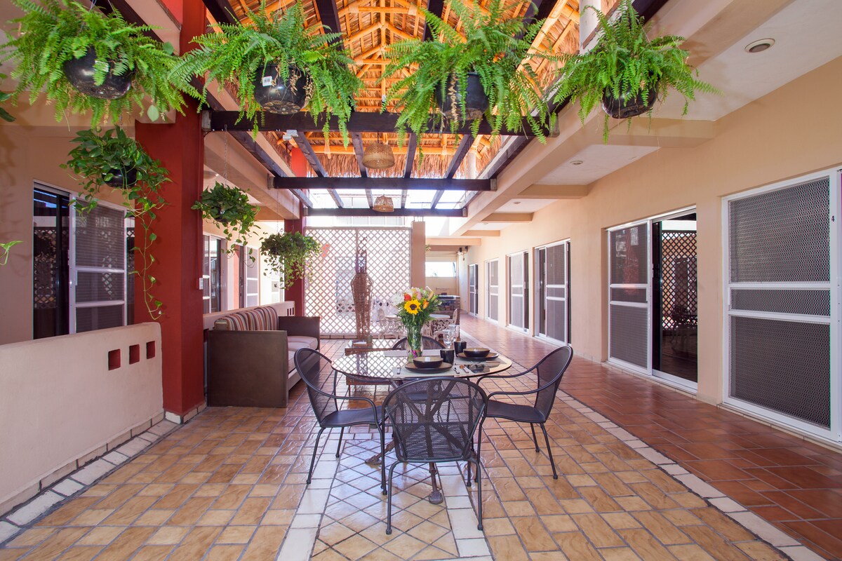 A communal terrace area features a dining table set for four, surrounded by potted plants hanging from the ceiling. Large windows provide natural light, and a comfortable seating area is visible in the corner. The flooring is designed with a patterned tiled layout.