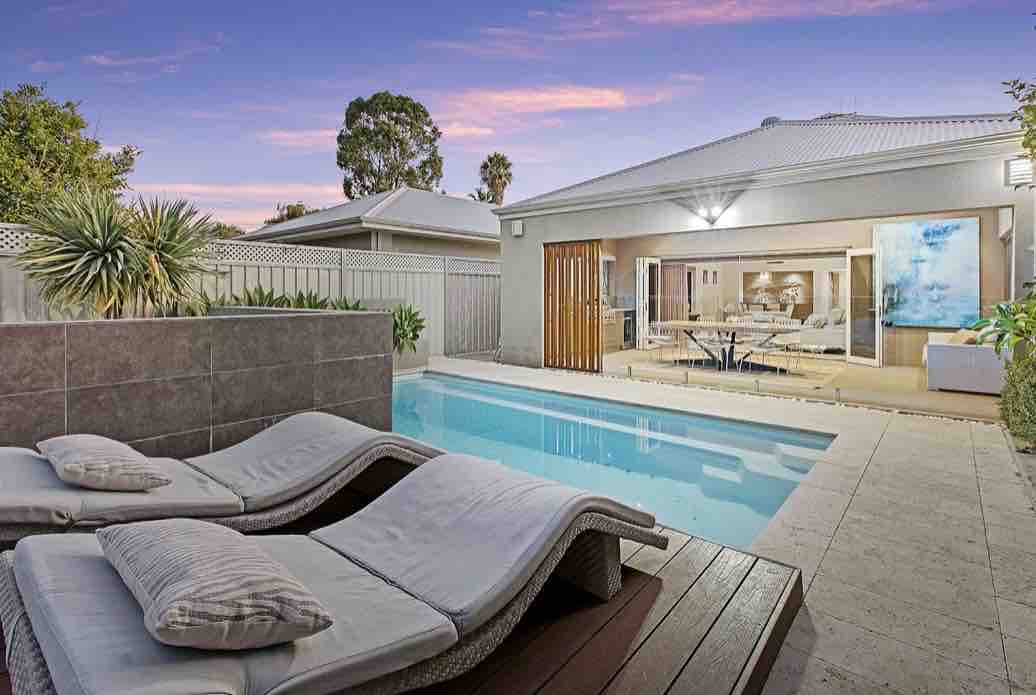 A private pool area is framed by a sleek, modern design. Comfortable sun lounges are positioned along the wooden deck beside the clear water. Bi-fold doors lead to a spacious indoor entertainment area, seamlessly connecting the outdoor and indoor spaces.