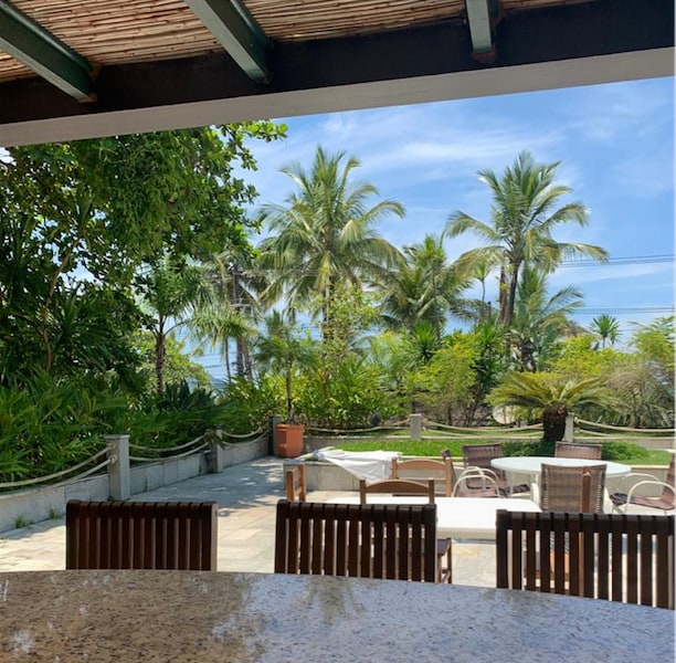 A view from the terrace reveals a spacious outdoor area surrounded by lush tropical plants. Glass tables and comfortable chairs are positioned for relaxation, with the bright blue ocean visible in the background under a clear sky.