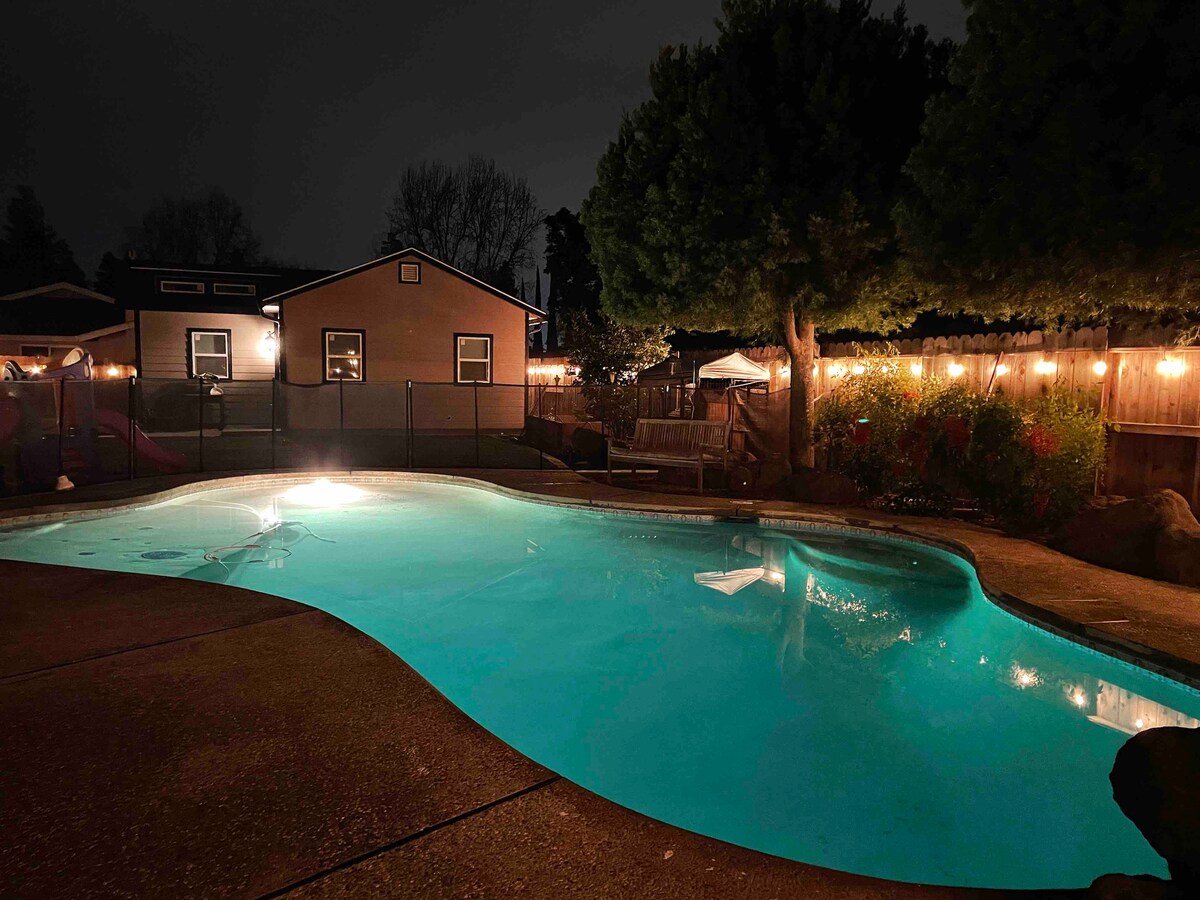 The illuminated pool features tranquil turquoise water under nighttime lighting. Surrounding the pool, string lights enhance the atmosphere, while a well-maintained yard is accented by mature trees and a fence. The background includes a guest house and outdoor seating areas.
