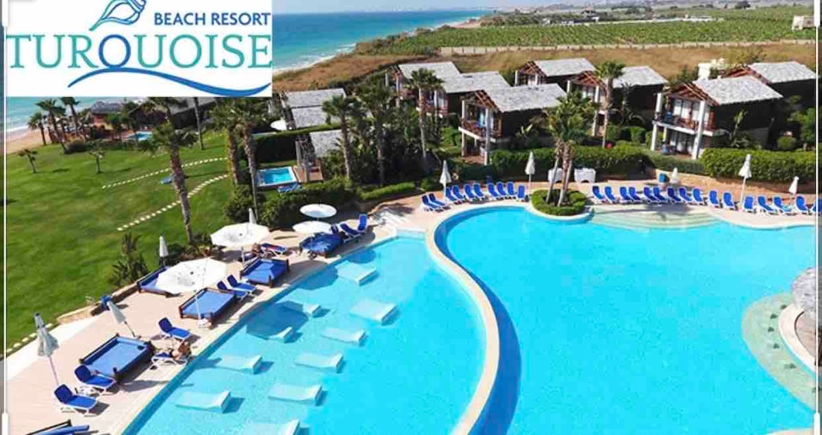 An aerial view showcases a large swimming pool surrounded by lounge chairs, with an expansive green lawn and palm trees visible nearby. The turquoise water of the pool contrasts with the sandy beach in the background, enhancing the serene coastal atmosphere.