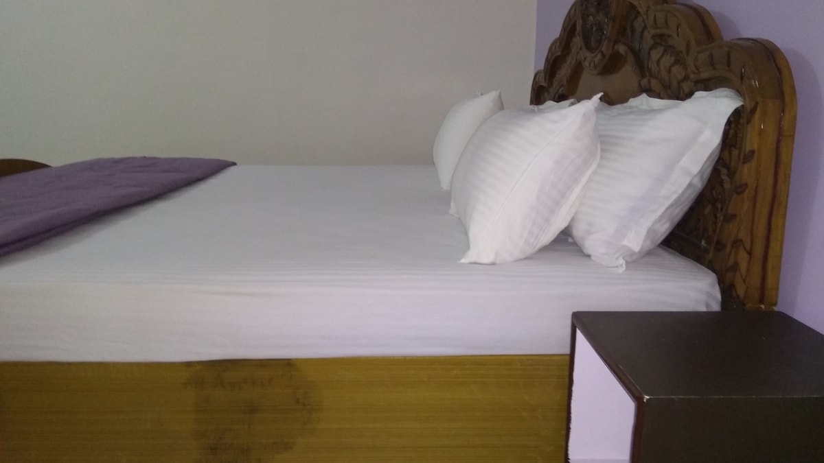 A spacious bed with a carved wooden headboard is complemented by white pillows and crisp bedding. A bedside table is positioned beside the bed, providing a functional space for personal items.
