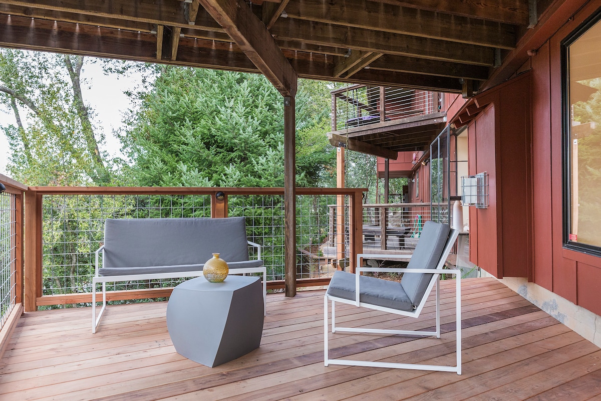 An outdoor deck features comfortable seating arrangements, including a grey chair and a sleek bench. A small, modern table is situated between them. Lush greenery is visible in the background, creating a serene outdoor environment.