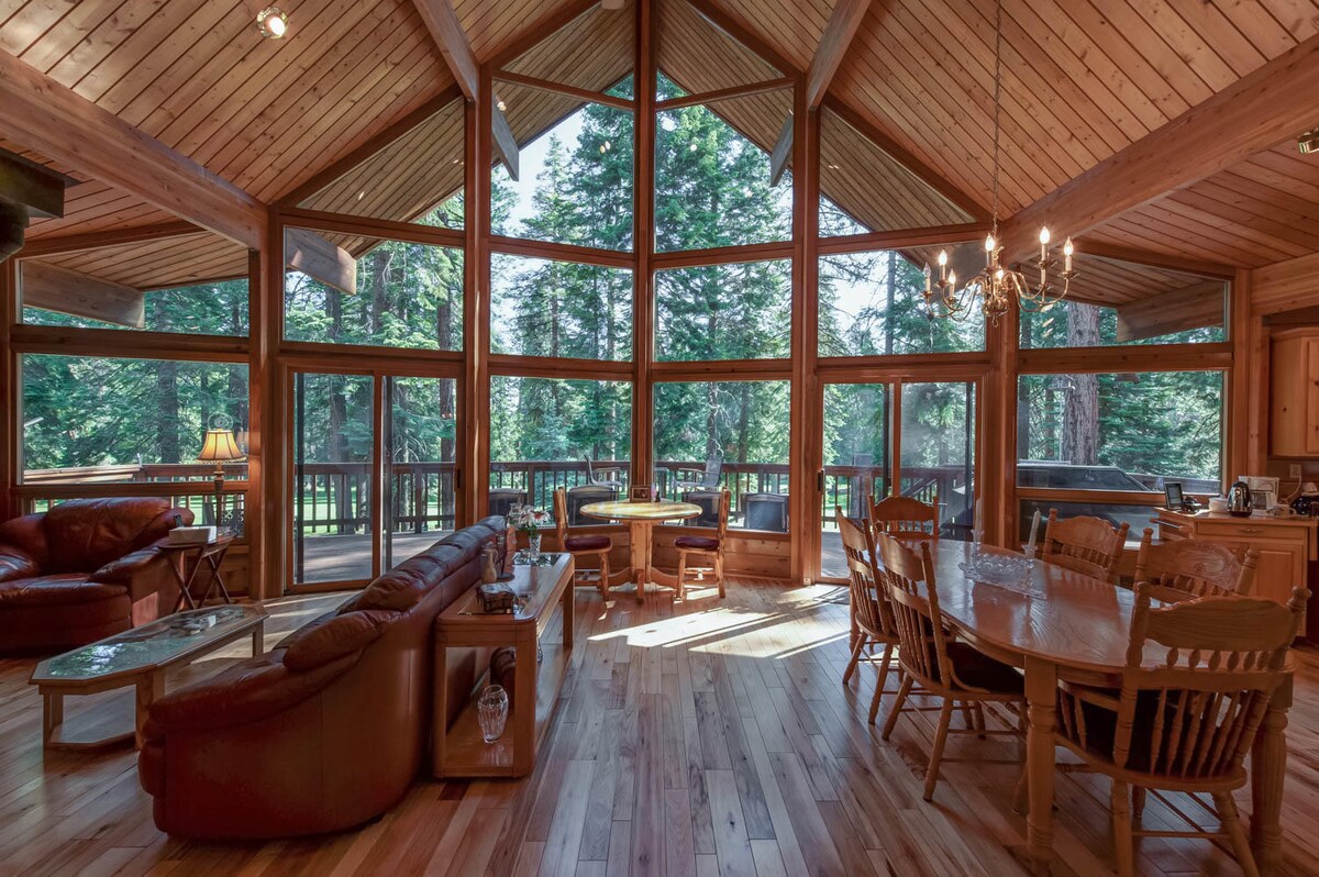 A spacious living area features large windows that invite abundant natural light and offer views of the surrounding trees. A wooden dining table is accompanied by several chairs, while a comfortable leather sofa and coffee table provide a cozy seating area. Wood flooring complements the warm wood accents.