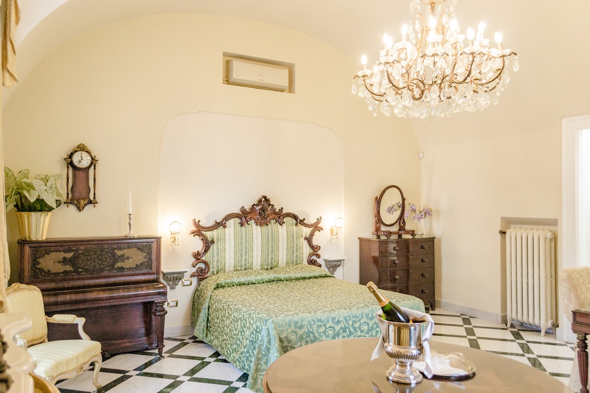 A spacious room features an elegant green-patterned bedspread on a large bed, complemented by vintage furnishings including a piano and a dark wood dresser. A chandelier hangs above, providing soft lighting, while marble flooring adds a classic touch to the ambiance.
