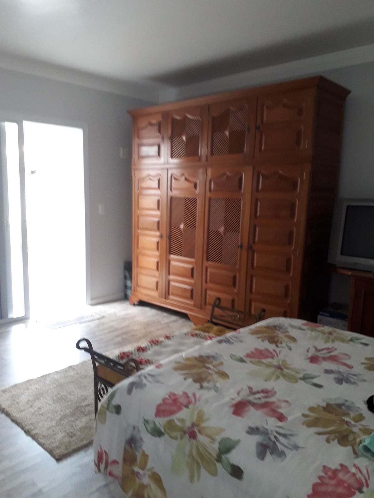 A well-lit bedroom features a spacious bed draped with a floral-patterned blanket. A large wooden wardrobe stands against one wall, while a television is positioned on a wooden stand nearby. Natural light enters through a sliding door, illuminating the room.
