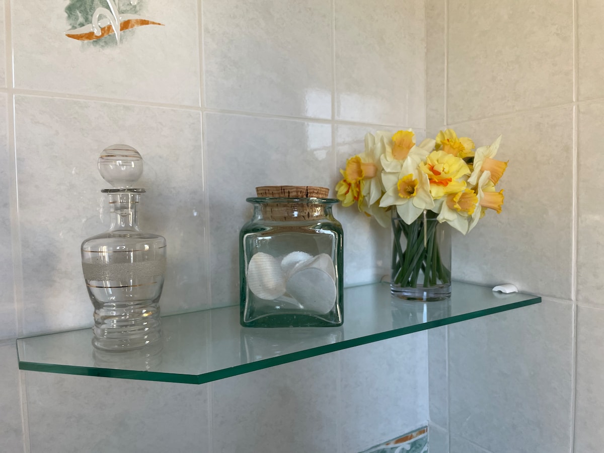 A glass shelf is shown, featuring a decorative clear bottle, a jar containing shells, and a vase filled with yellow daffodils. The soft light reflects gently on the smooth surface, creating a calm and fresh atmosphere.