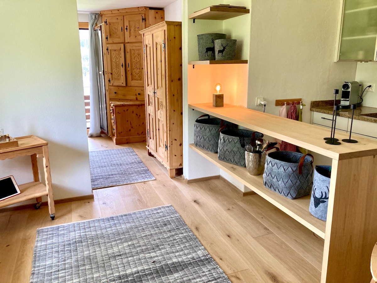 A warm entrance area features light wooden shelving displaying decorative baskets and a lamp. Natural light enters through an open door leading to a balcony. Soft area rugs are placed on the wooden floor, enhancing the inviting ambiance of the space.