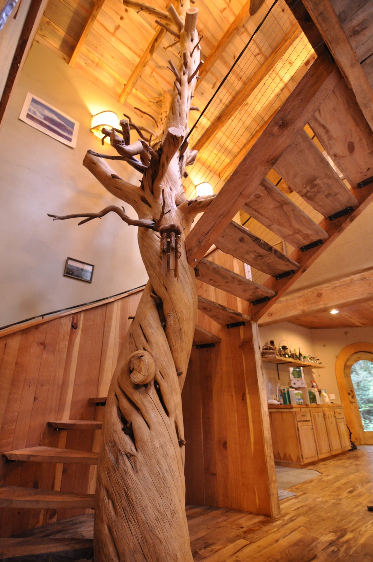 A unique spiral staircase features a stunning, twisted juniper tree trunk as a centerpiece, connecting two levels of the home. The surrounding wooden walls and floors create a natural atmosphere, complemented by soft lighting overhead, enhancing the artistic design of the space.