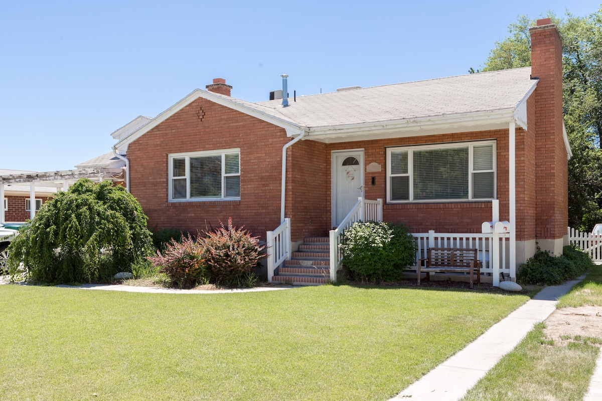A red brick exterior is featured on this single-story home, highlighted by a well-maintained lawn. A set of steps leads to the front door, flanked by large windows for natural light. A wooden bench is positioned on the porch, adding to the welcoming ambiance.