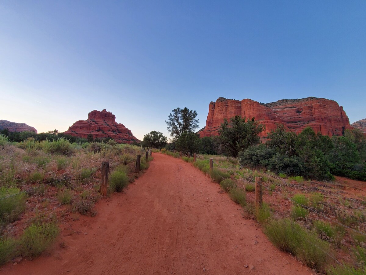 A sandy path winds through a landscape of red rock formations and green vegetation. The vibrant colors of the rocks are highlighted by the soft glow of evening light, creating a serene atmosphere inviting exploration.