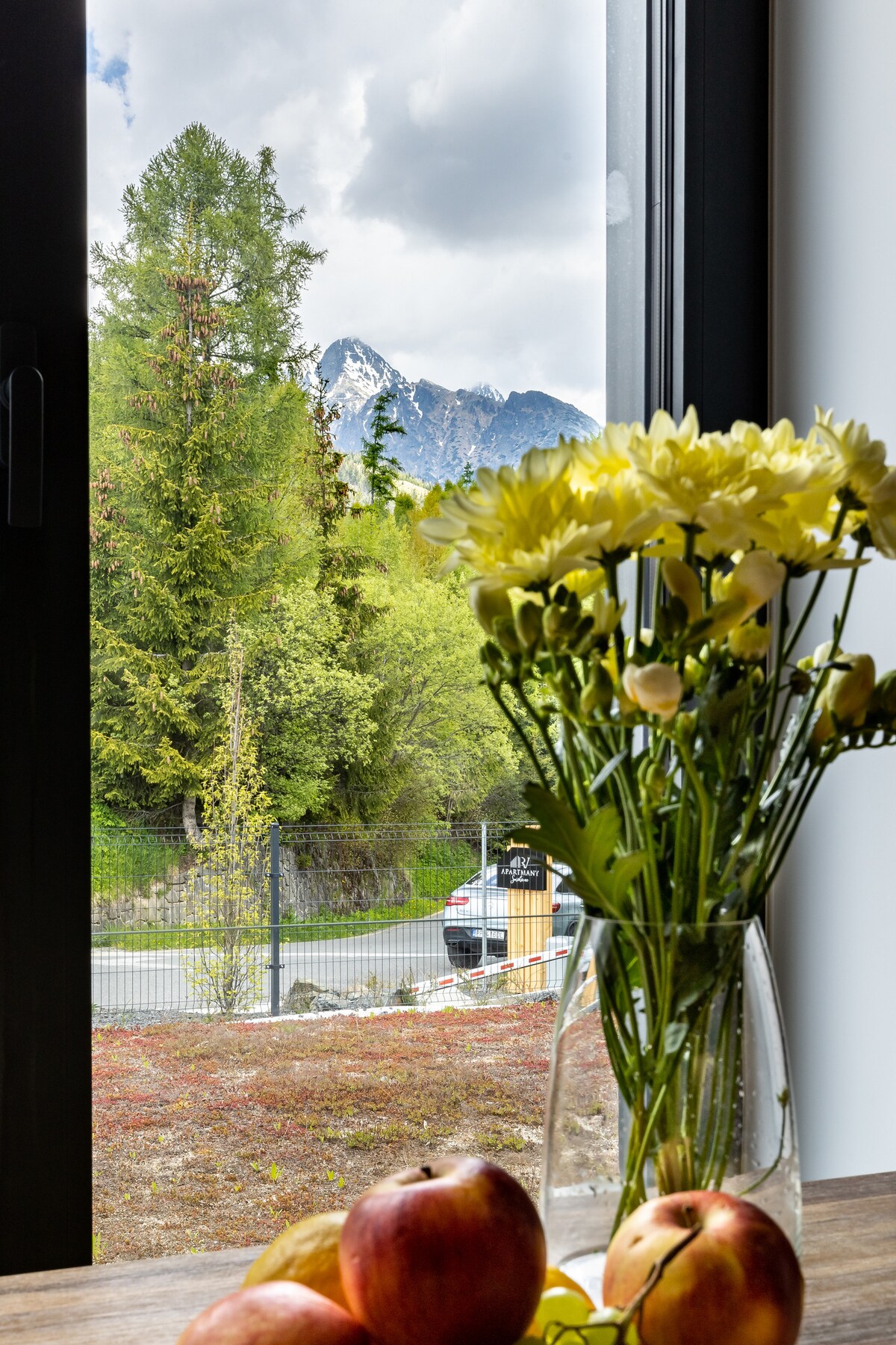 A view from the window features a mountain landscape framed by trees. A vase of flowers and a bowl of apples are placed on a table in the foreground, creating a serene atmosphere.