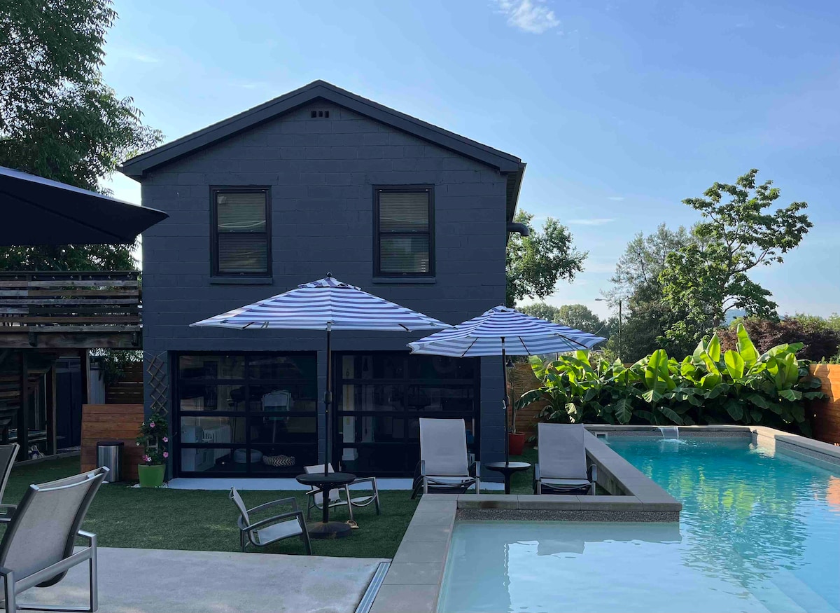 A modern gray cottage is showcased, complemented by a private pool area. Two striped umbrellas provide shade over lounge chairs arranged neatly beside the sparkling water. Lush greenery surrounds the space, enhancing the relaxed ambiance of the outdoor setting.
