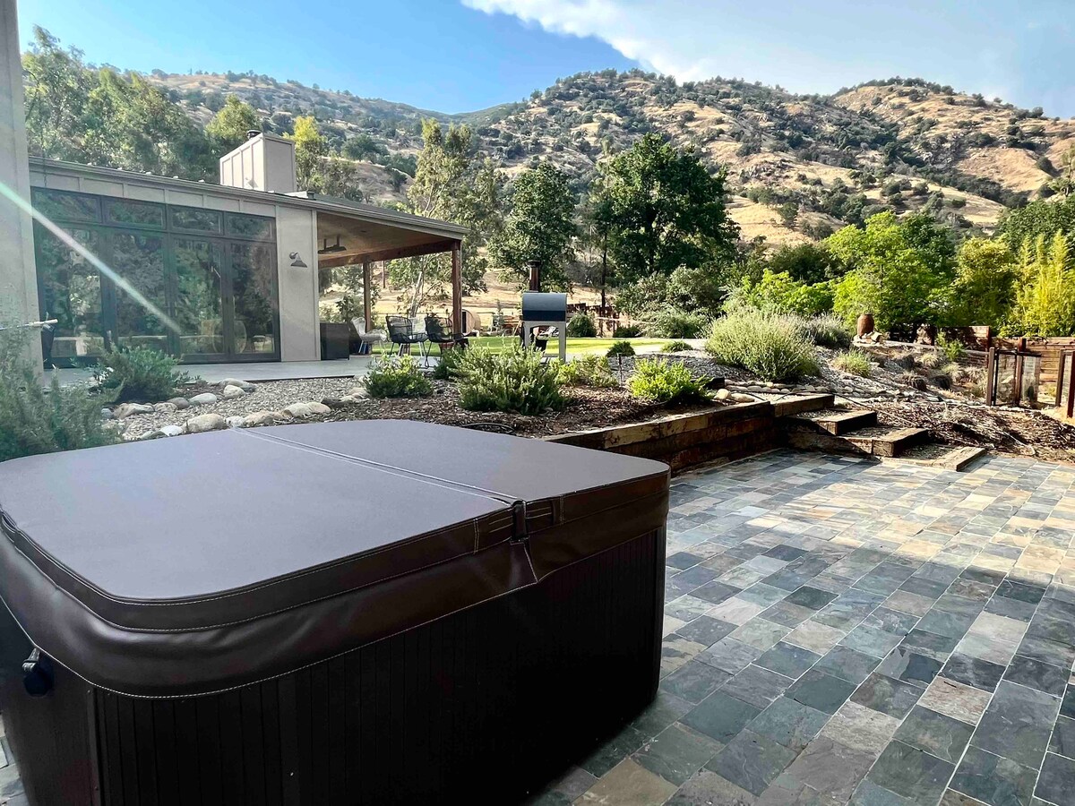 A hot tub is located on a stone patio surrounded by landscaped gardens, with rolling hills in the background. Large glass doors provide a view of the modern interior, seamlessly merging with the outdoor space. Lush greenery and seating areas enhance the tranquil setting.