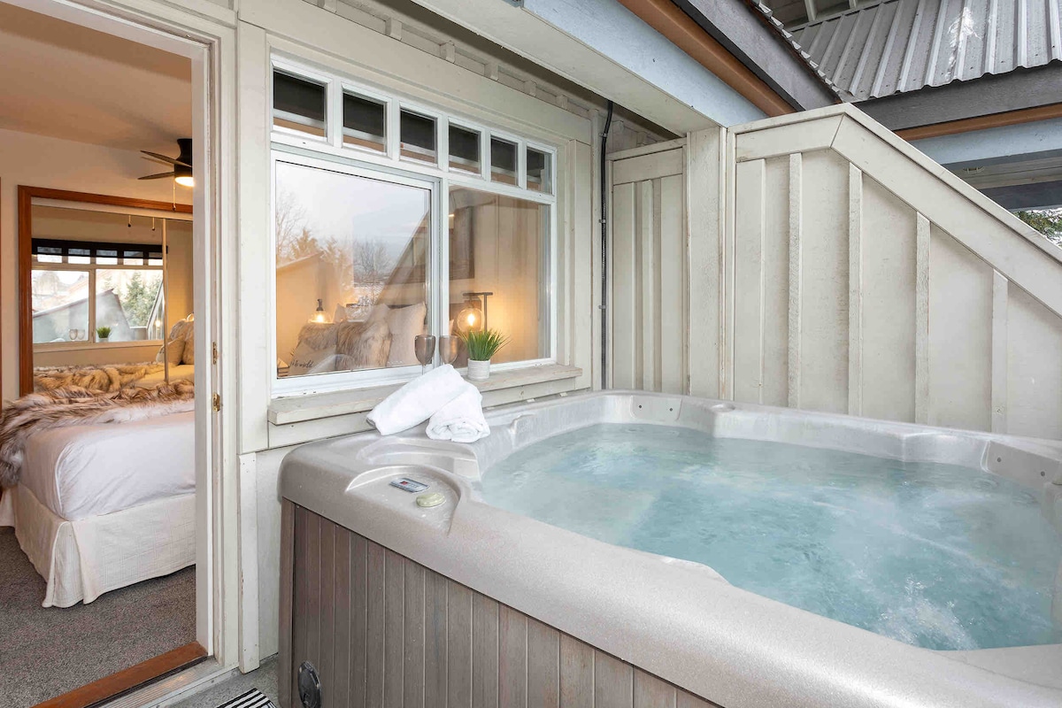An inviting private hot tub is featured on a deck, surrounded by rustic wooden walls. A glimpse of a cozy bedroom is visible through a large window, showcasing soft bedding and warm lighting. The scene emphasizes relaxation and comfort.