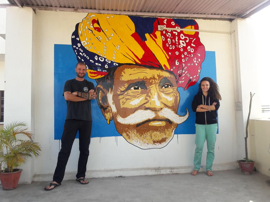 A vibrant mural featuring an abstract representation of an elephant is positioned on a plain wall, providing a striking backdrop. Two individuals stand at either side, adding a human element to the scene. Potted plants sit on the ground, enhancing the outdoor space.
