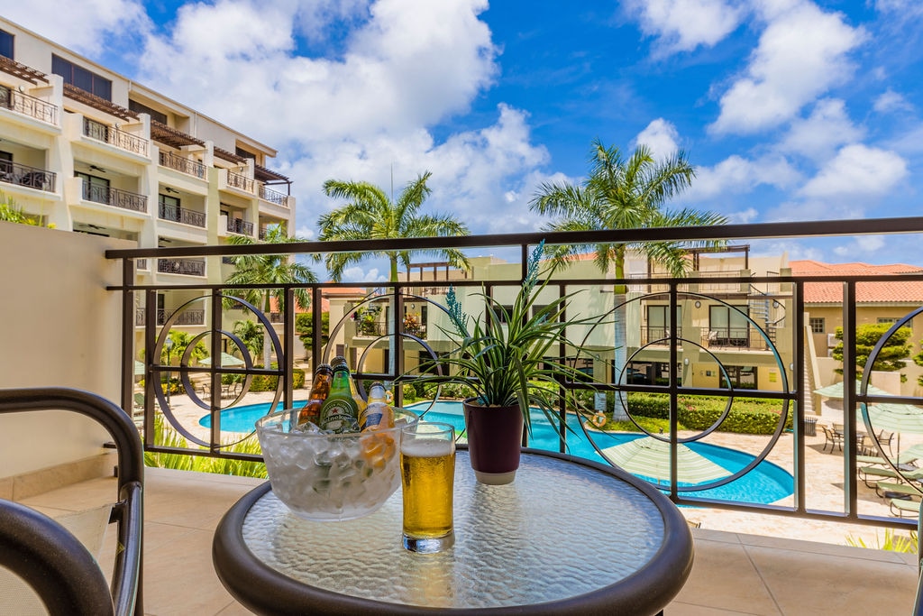A spacious balcony features a round glass-top table with two drinks and a small potted plant. The view overlooks a sparkling pool surrounded by well-kept greenery and palm trees, with the building's exterior in the background under a bright blue sky.