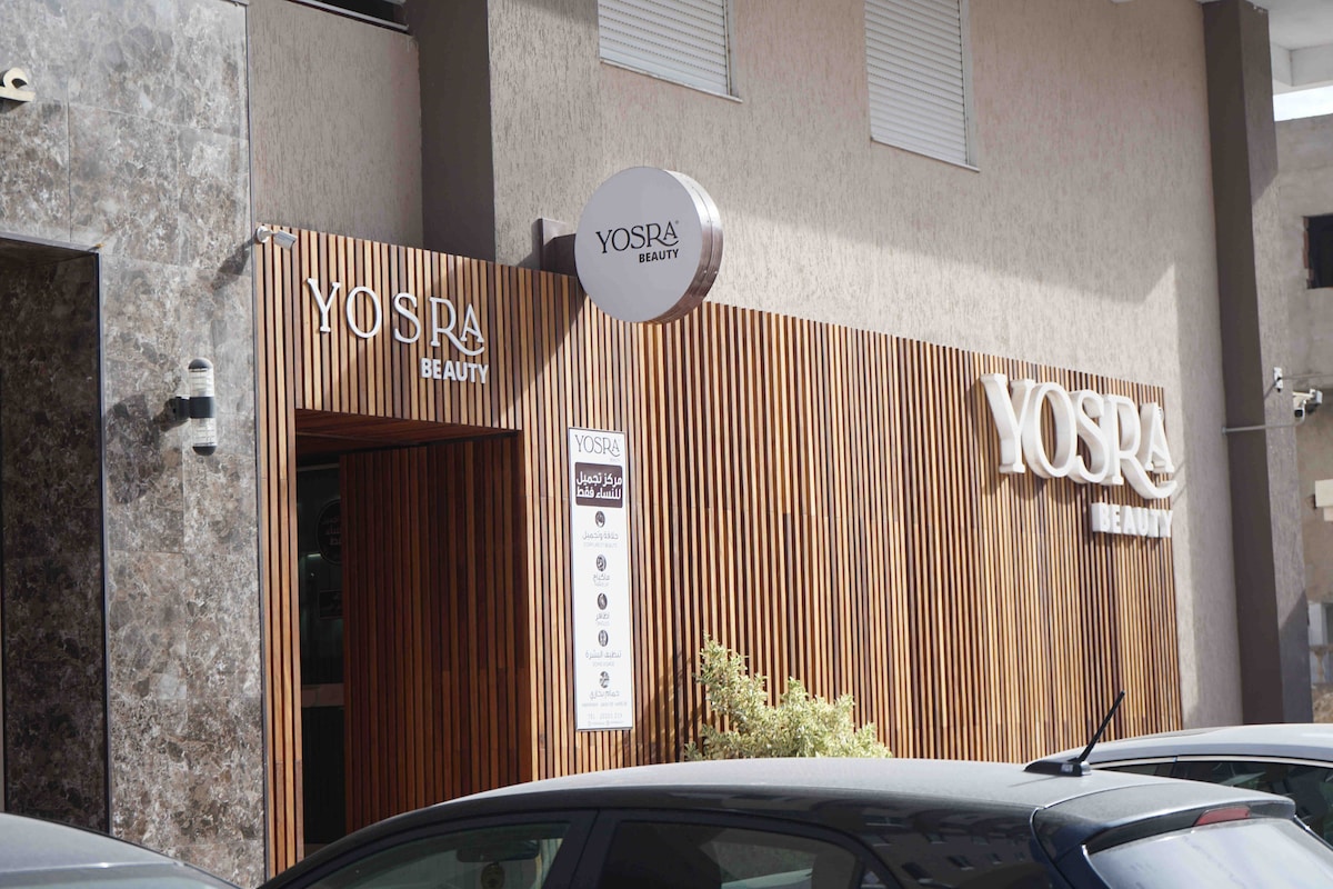 An exterior view of a beauty establishment is presented, featuring wooden paneling and a modern sign displaying the name 'Yosra Beauty.' The entrance is highlighted by a circular sign above the door, creating a welcoming appearance. Surrounding greenery adds a touch of nature.