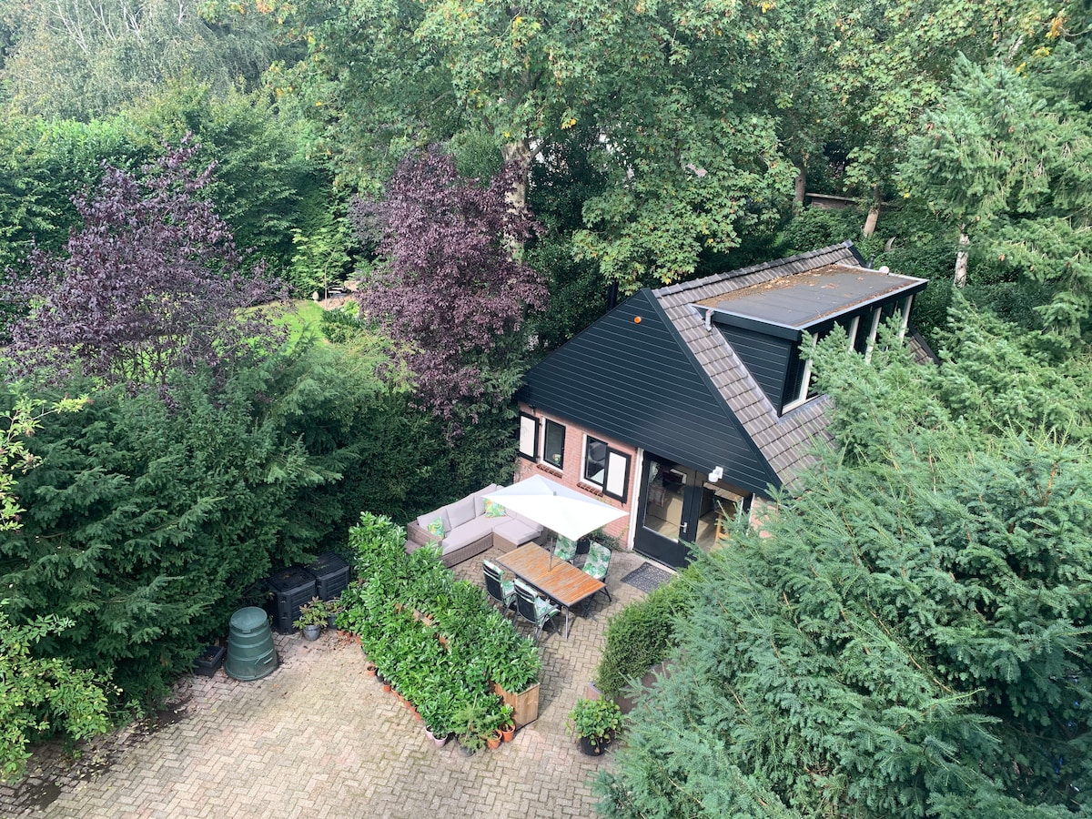 An aerial view showcases the charming Garden House nestled among lush greenery. A spacious outdoor dining area, featuring a wooden table and chairs, is visible on the terrace. Surrounding foliage includes tall trees and vibrant bushes, offering a private and tranquil atmosphere.
