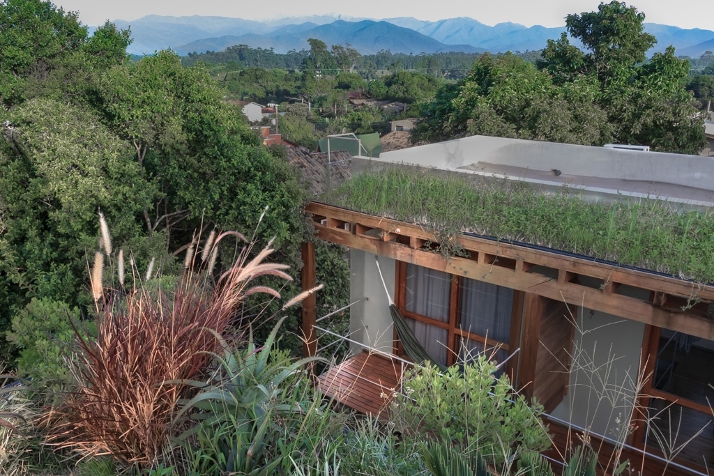 The image displays a modern duplex with a green rooftop, surrounded by lush vegetation. The structure is partially obscured by plants, and the background features rolling mountains under a clear sky, emphasizing the natural setting.