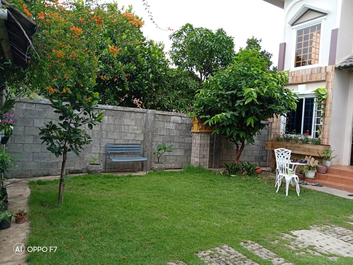A spacious outdoor area is depicted, featuring a well-maintained lawn surrounded by lush greenery. A blue bench is positioned on the left side, while white chairs and a small table are located on the right. Potted plants add greenery along the wall, enhancing the inviting atmosphere.