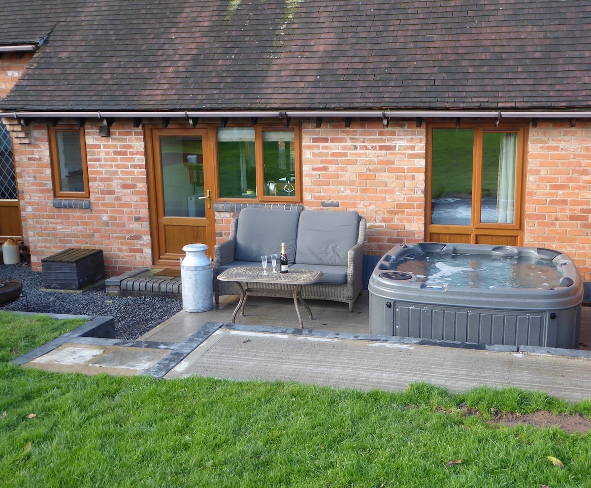 An outdoor area features a cozy seating arrangement with a grey sofa and a small table paired with complimentary drinks. Next to the seating, a salt water hot tub is displayed, inviting relaxation. The space is framed by a well-maintained lawn and the brick exterior of the Coach House.