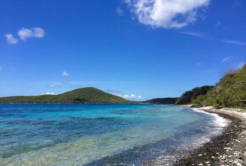 A serene coastal view showcases clear turquoise waters lapping against a rocky shore. Lush green hills rise gently in the background under a bright blue sky dotted with a few white clouds, creating a sense of tranquility and connection with nature.